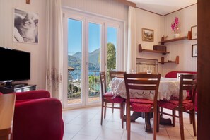 A room with a TV, red chairs, a dining table with lace tablecloths, and a balcony with a mountain view.