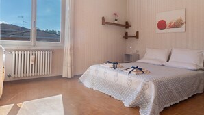Een slaapkamer met een groot raam, een radiator, een bed met een beddeken met een patroon, en een wandplank met een plant.