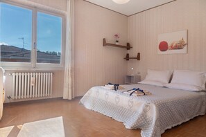 Quarto com uma janela grande, um radiador, uma cama com colcha estampada e uma prateleira na parede com uma planta.