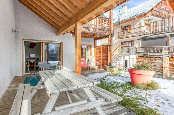 Una terraza de madera con mobiliario exterior, una zona techada y una escalera que lleva a un balcón.