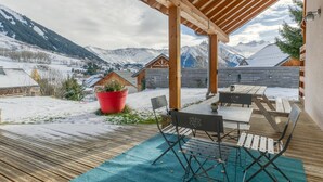 A covered outdoor area with a wooden deck, a table with chairs, and a view of snow-covered mountains.