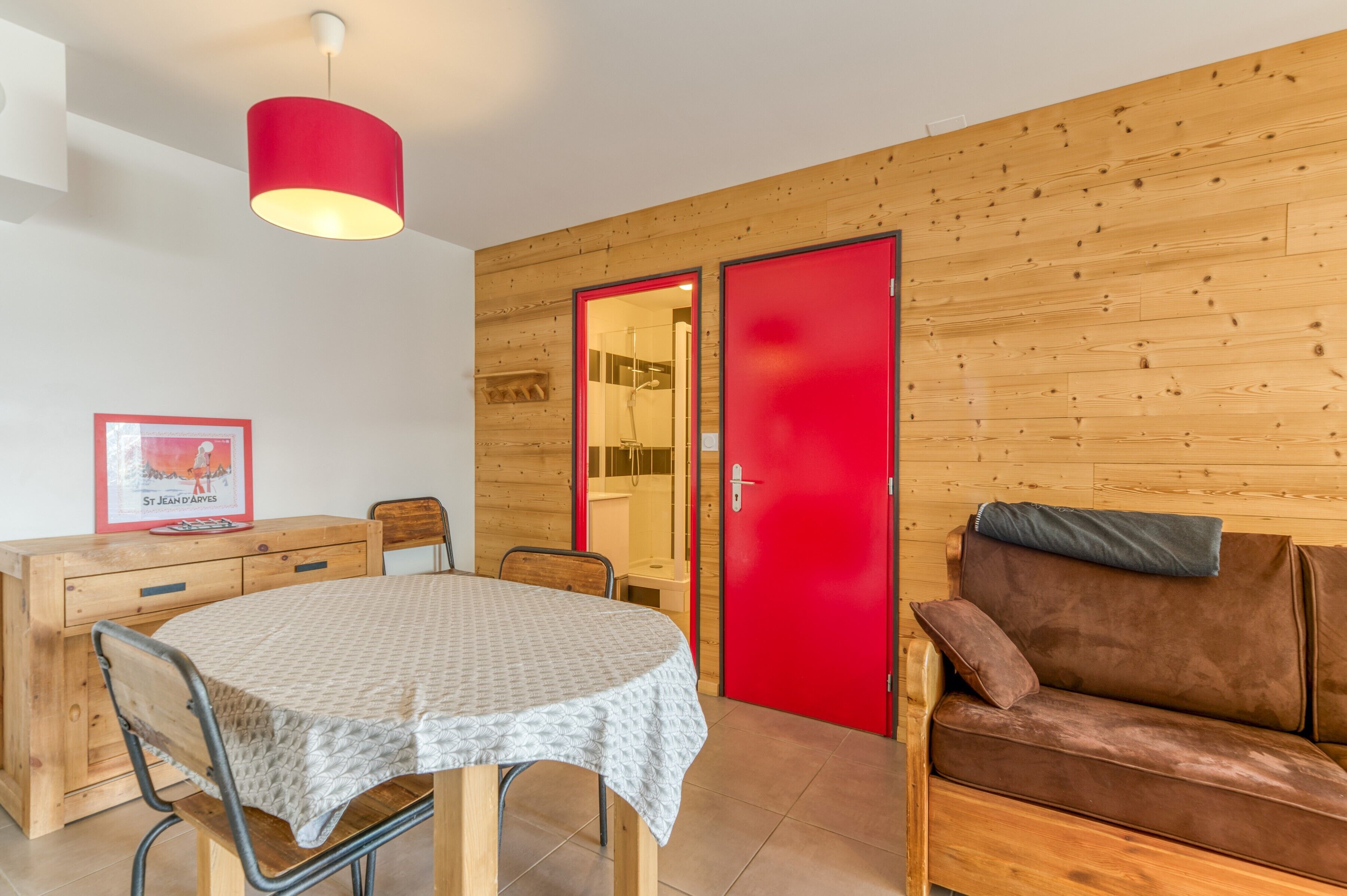 A room with a wooden wall, a red door, a dining table with chairs, and a brown sofa.