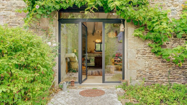 Un bâtiment en pierre avec une entrée dotée d’une porte en verre, un petit espace extérieur avec un paillasson et de la verdure tout autour.