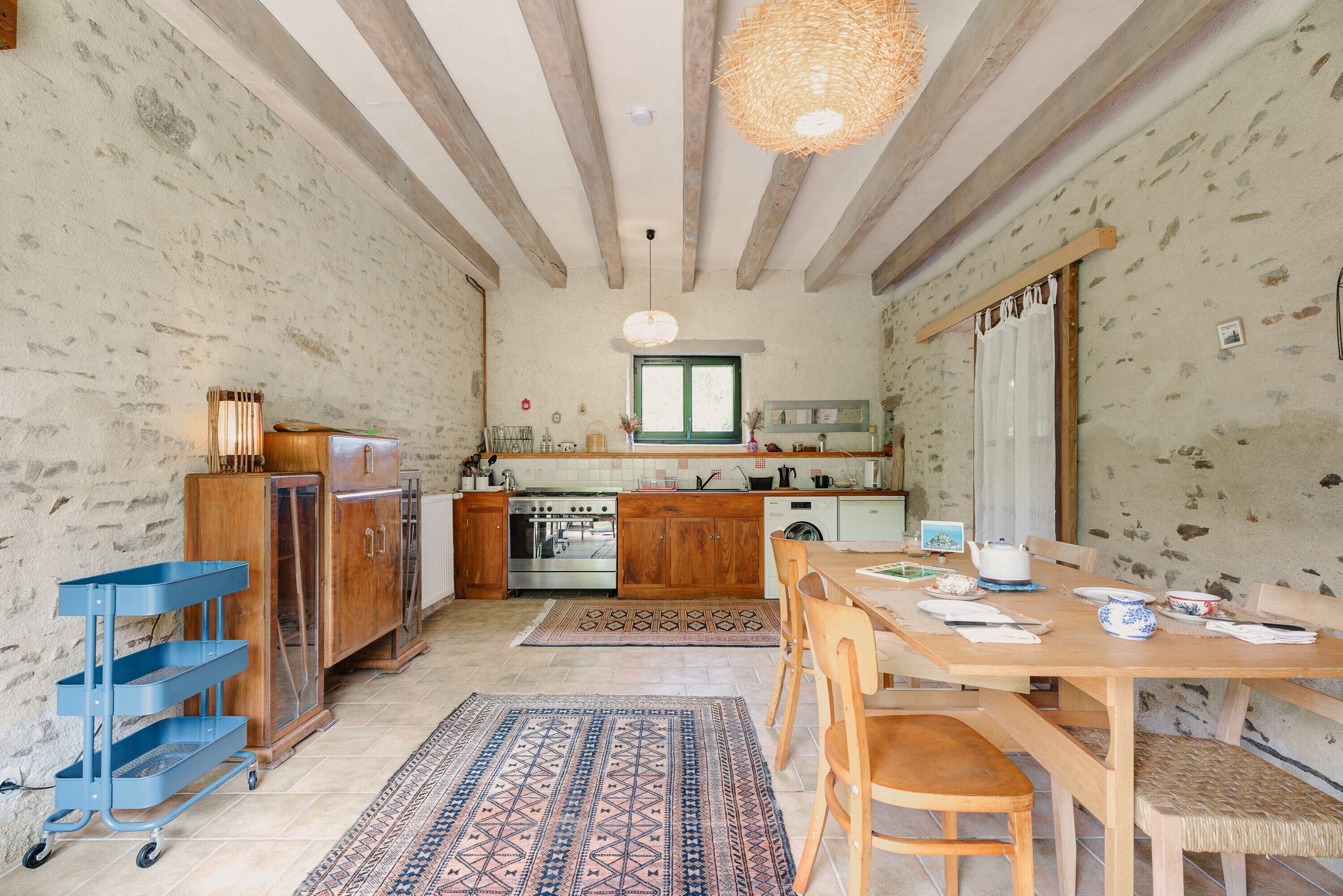 Une cuisine rustique avec des meubles de rangement en bois, une table à manger avec des chaises, un chariot bleu et un tapis à motifs.