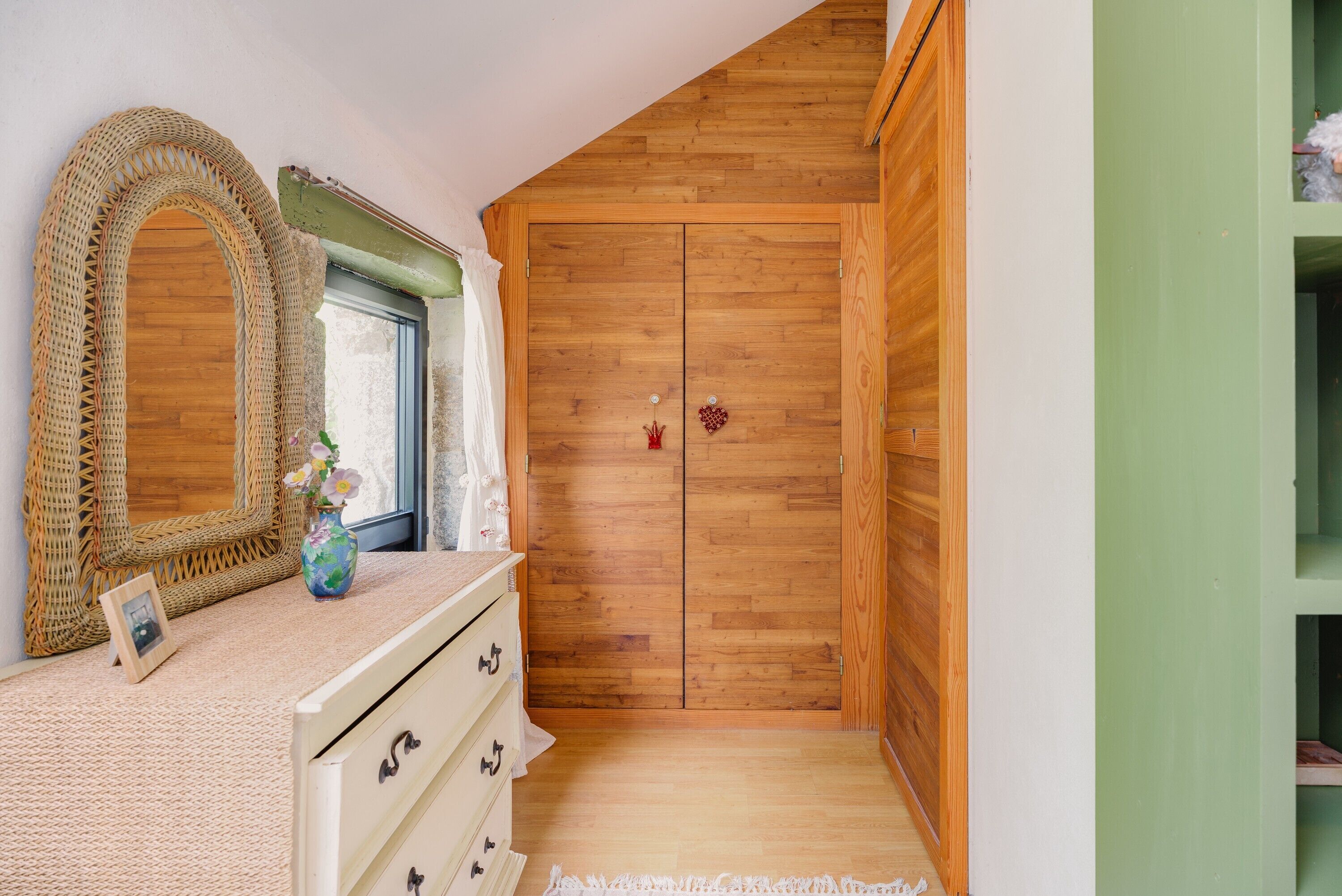 A room with a wooden wardrobe, a mirror, a dresser, and a vase with flowers.