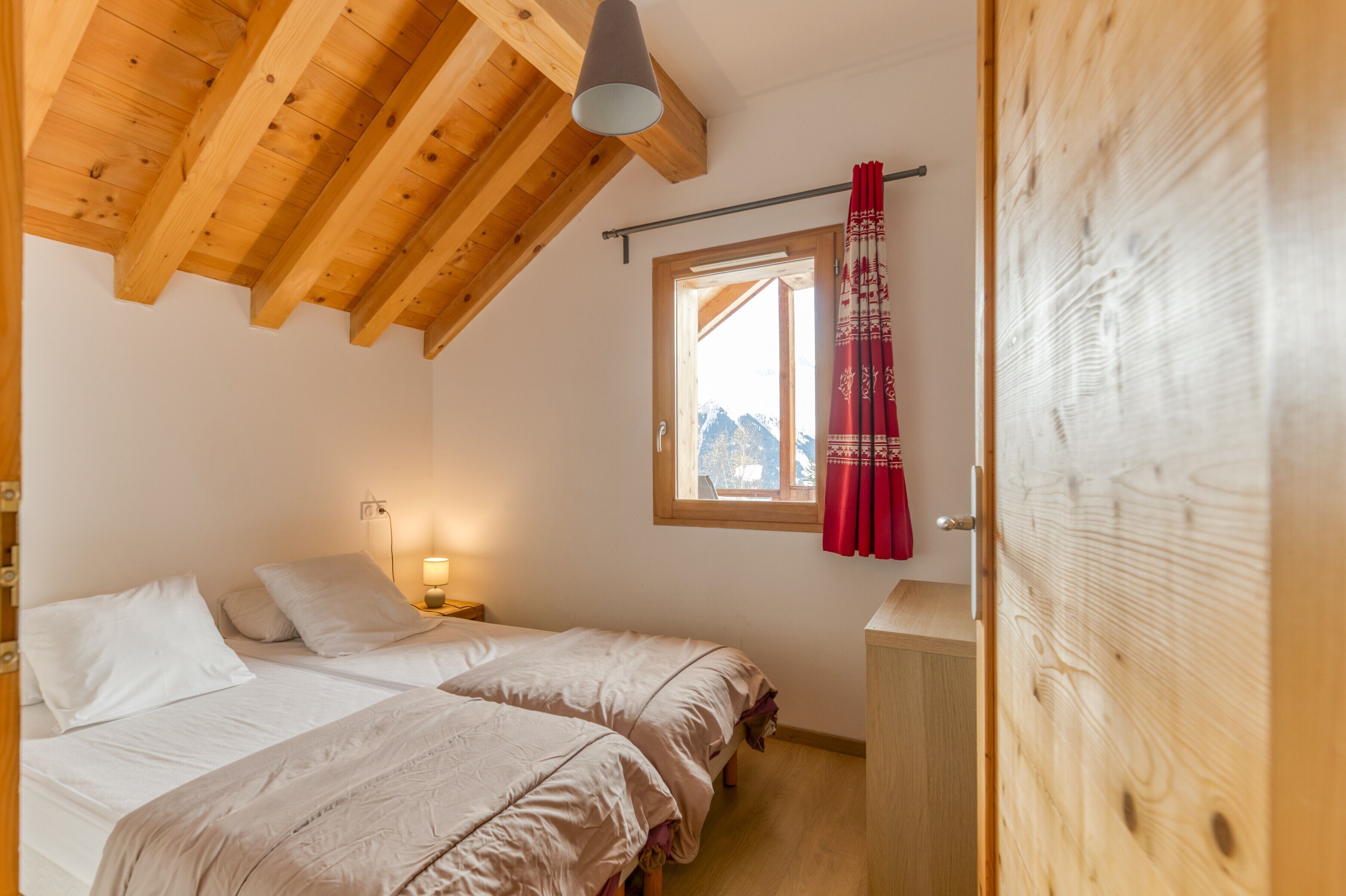 Een slaapkamer met twee bedden, houten balken in het plafond, een raam met uitzicht en een rood gordijn.