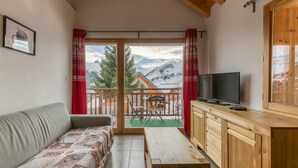 A cozy room with a sofa, a wooden table, a TV, and a balcony with a view of mountains.