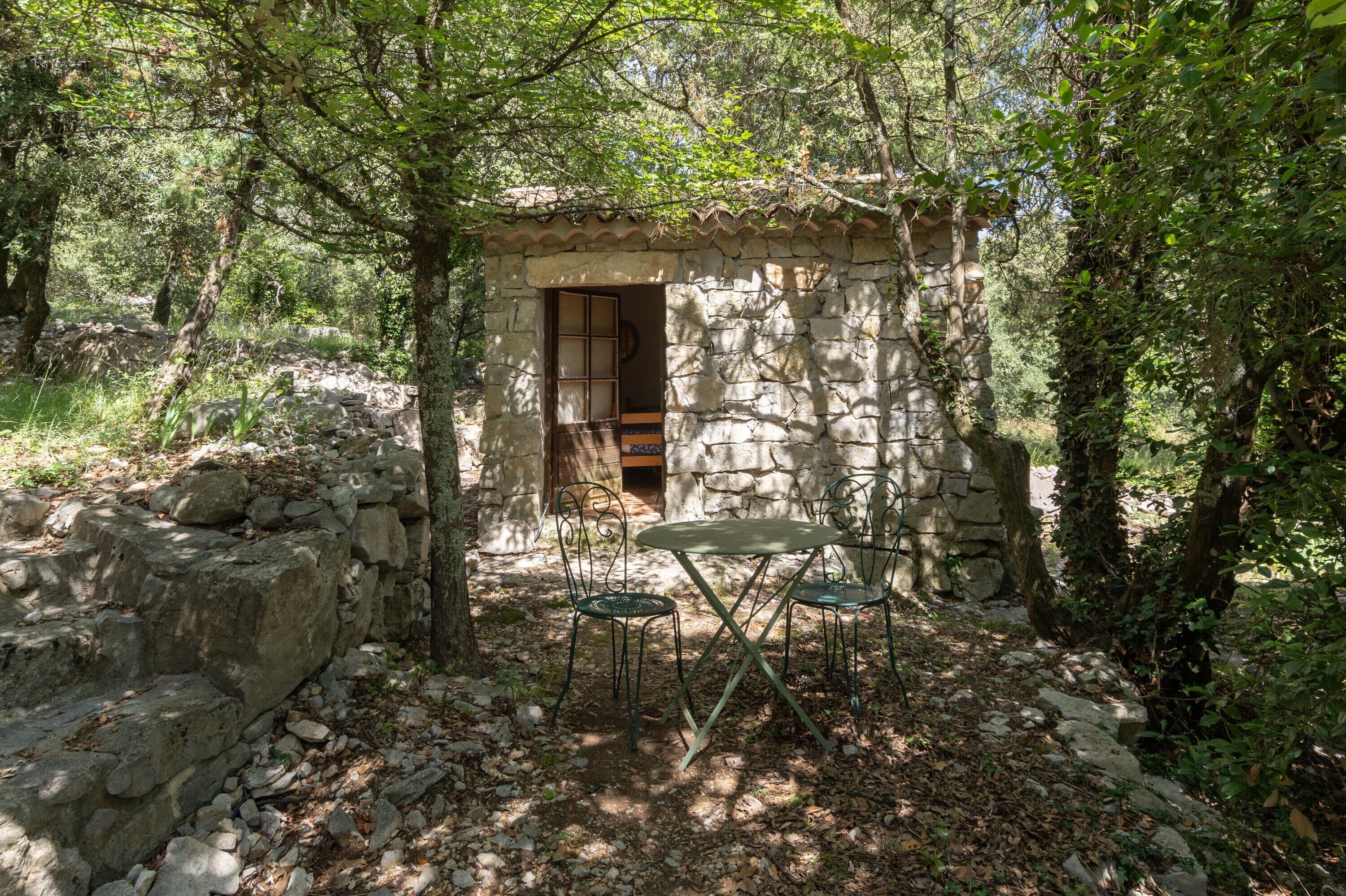 A rustic stone structure with a wooden door, surrounded by trees, and a small table with chairs in the foreground.