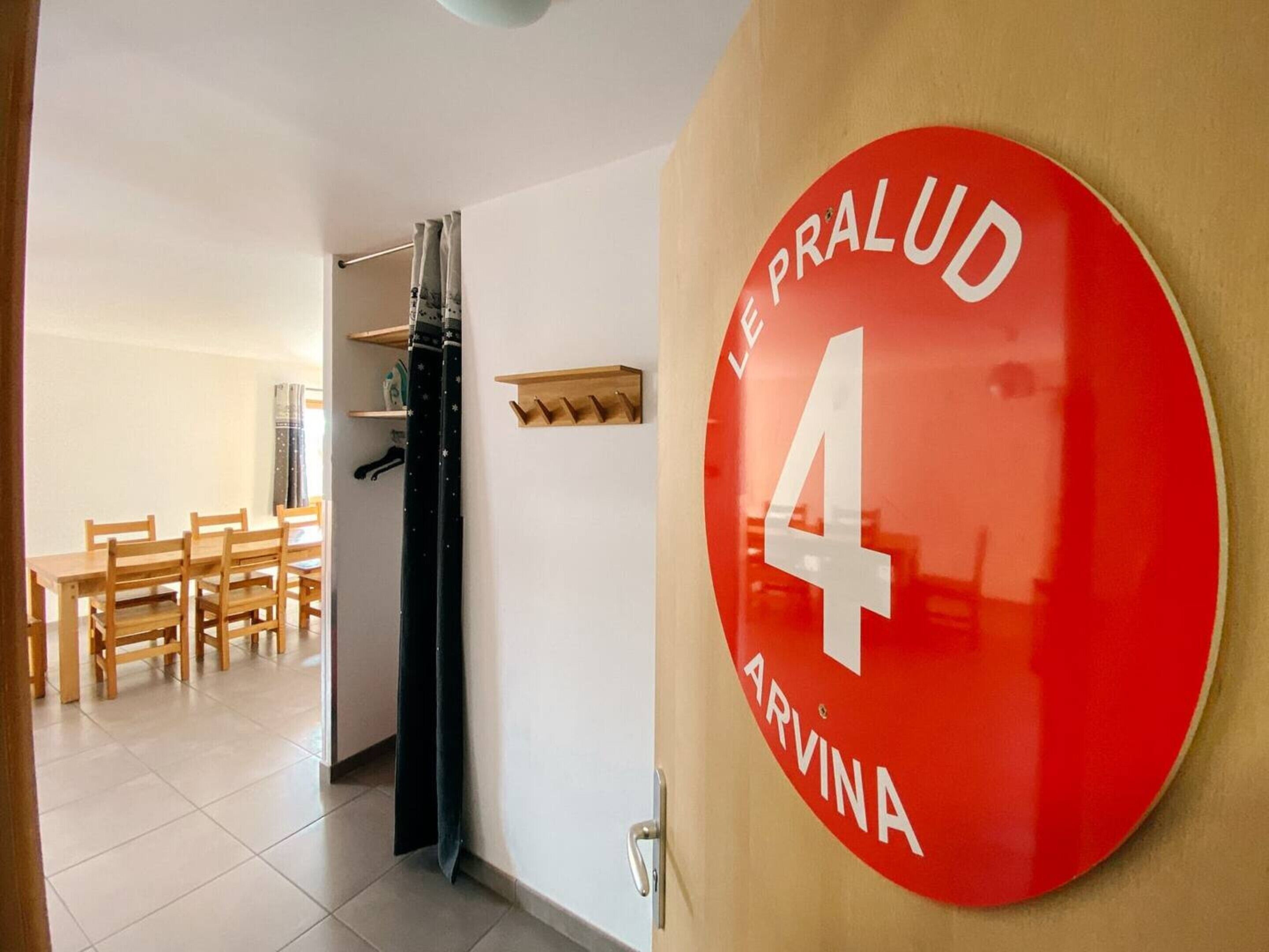 Een hal met een rood, rond bord met erop kamer 4, een houten tafel met stoelen, en een zwart gordijn.