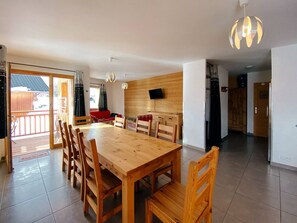 Un coin repas avec une table et des chaises en bois, un téléviseur fixé au mur et un balcon offrant une vue sur l’extérieur.