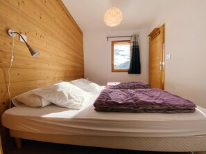 Une chambre avec un plafond en pente en bois, un lit avec une literie blanche et violette, un luminaire suspendu, une fenêtre avec des rideaux et une porte.