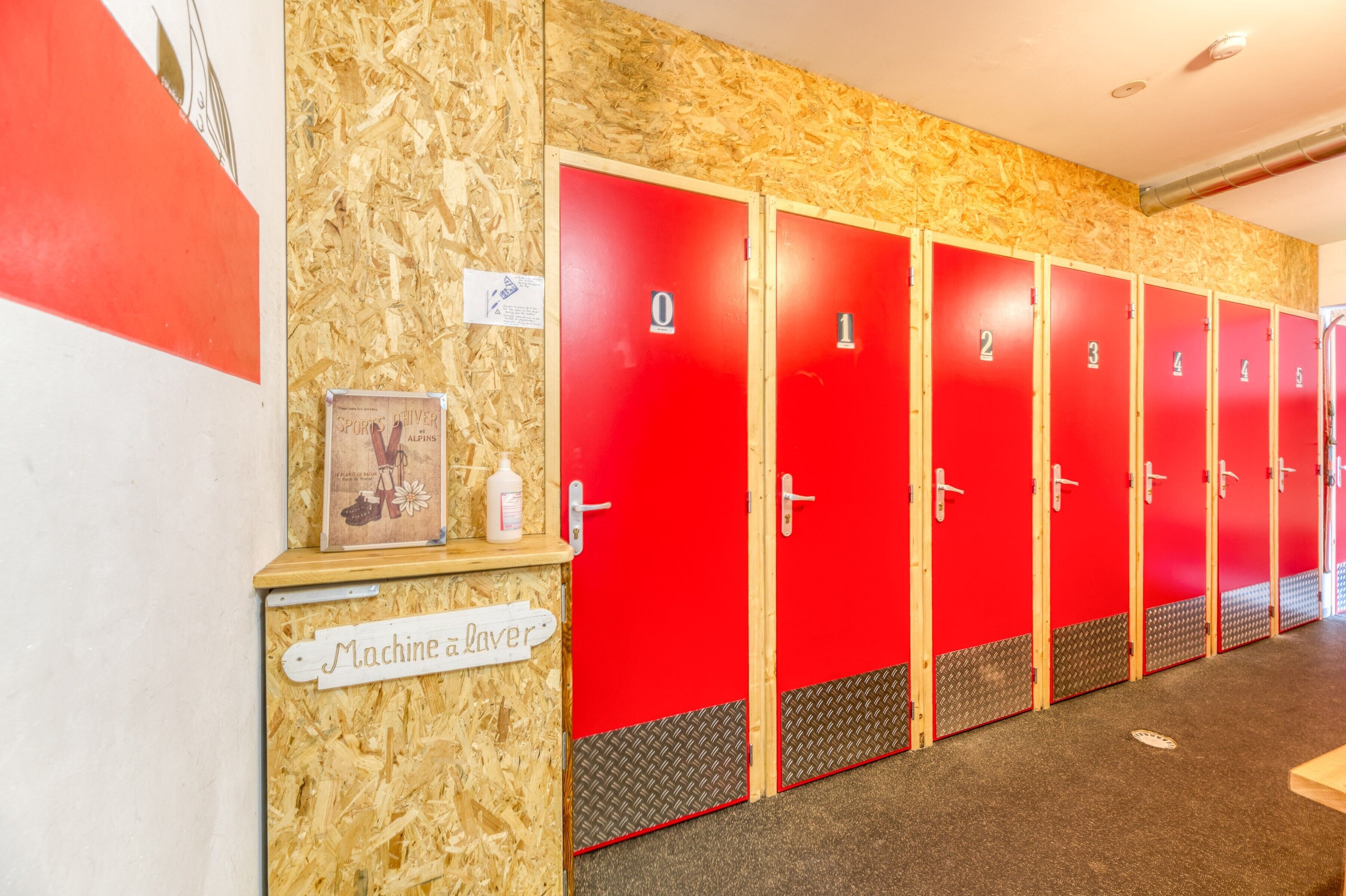 A row of red lockers with numbers 0 to 5, a wooden bulletin board with a sign reading 'Machine alaver,' and a textured wall.