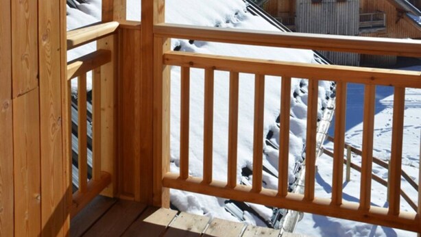 Un balcon en bois avec un luminaire en métal, une vue sur des montagnes enneigées et un chalet en bois en arrière-plan.