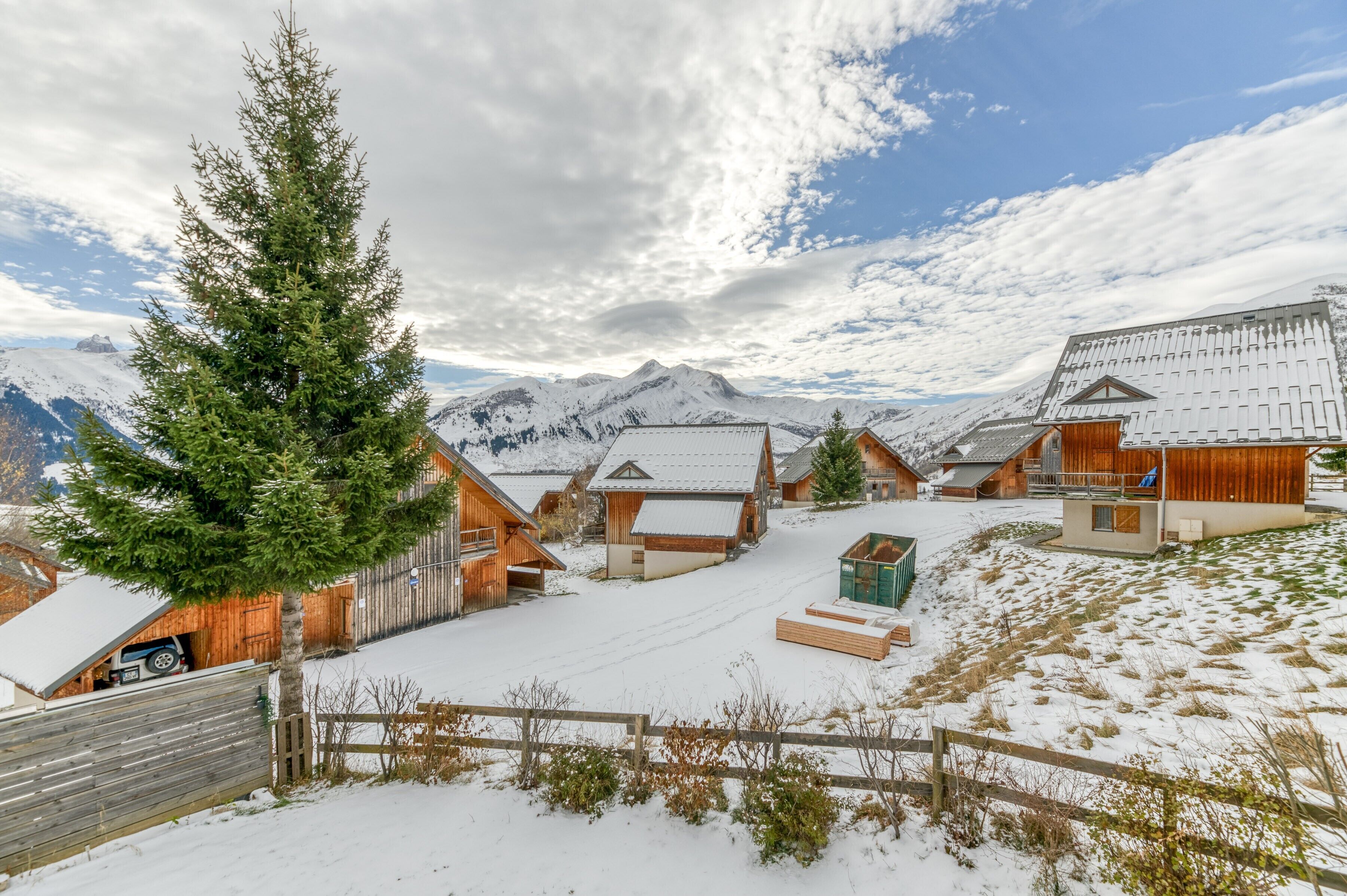 Ein verschneites Dorf mit Holzhäusern, eine große Nadelbaum und Berge im Hintergrund.