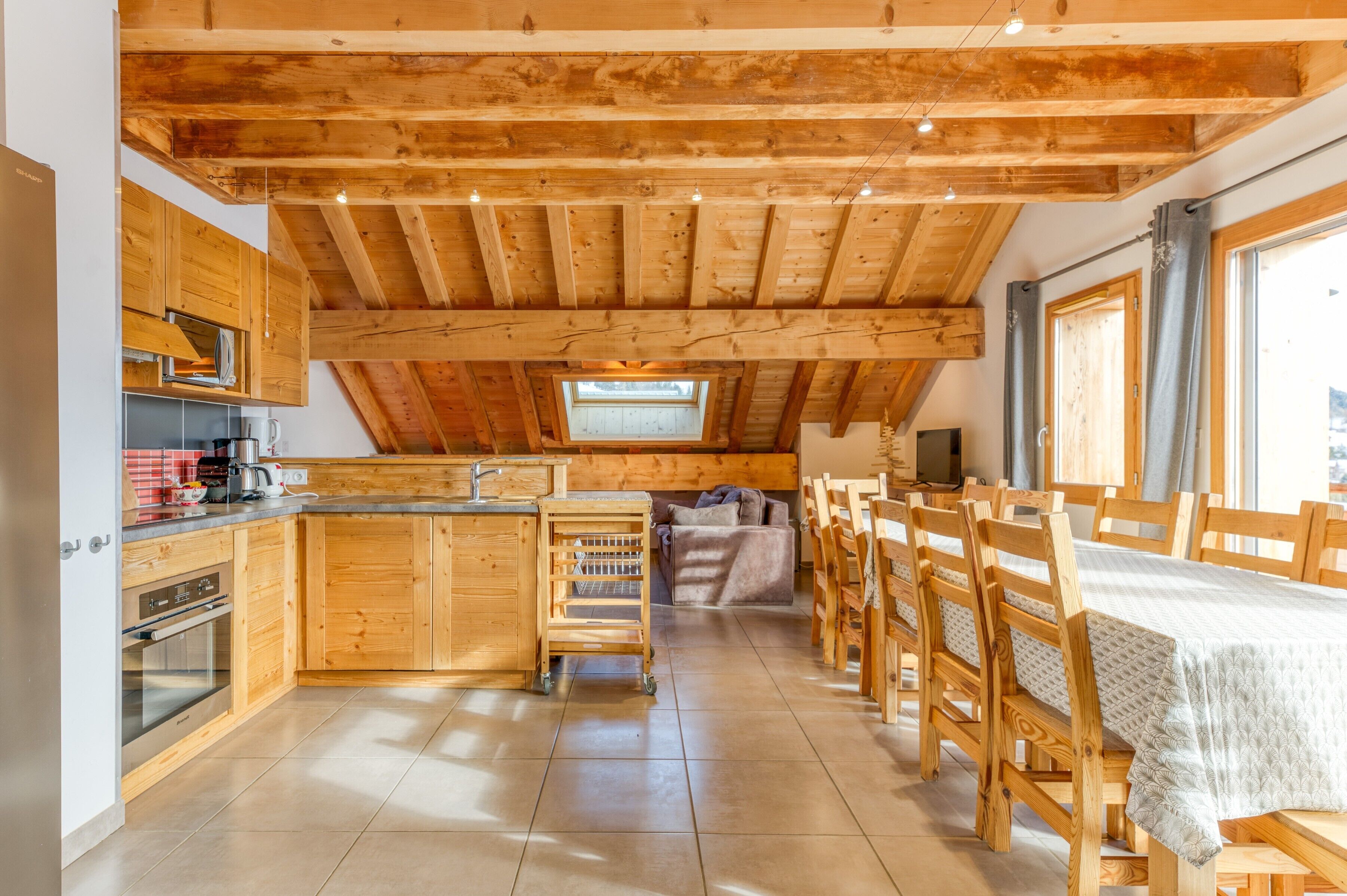 Een keuken met houten kasten, een eethoek met houten tafels en stoelen, en een schuine plafond met zichtbare balken.