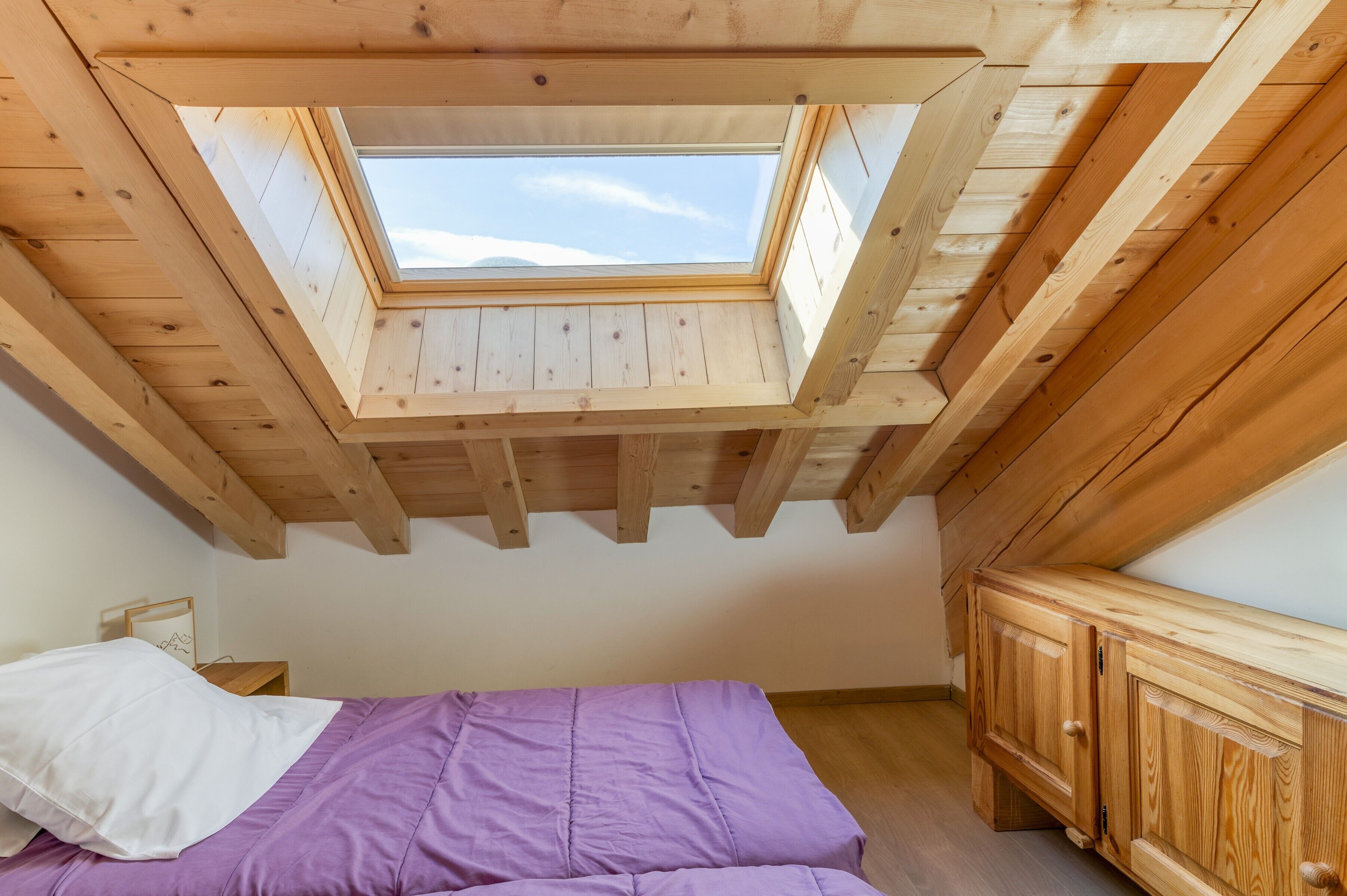 Une pièce avec un plafond en pente, une lucarne, un lit recouvert d’une courtepointe violette et des meubles en bois.