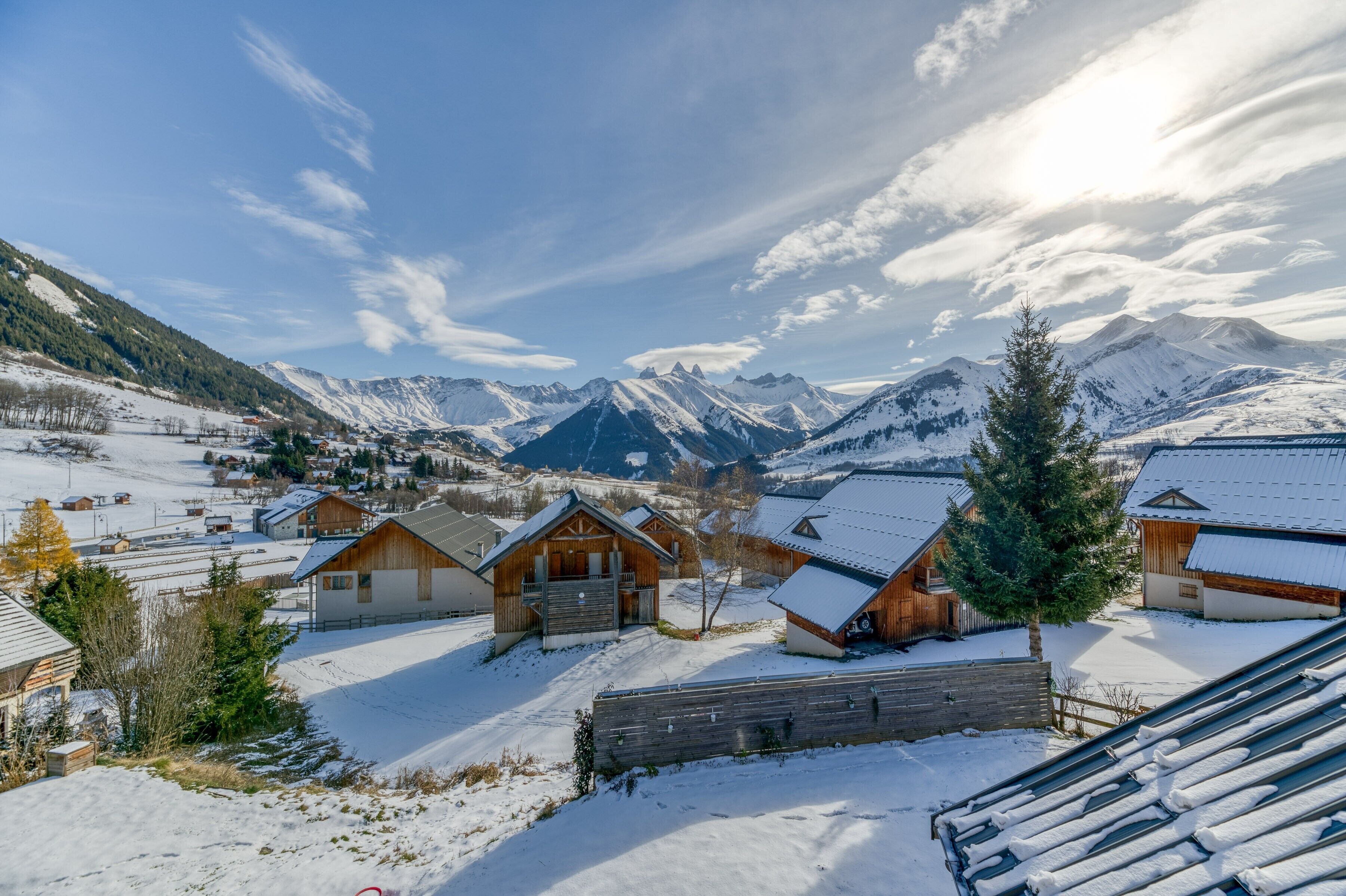 Un village enneigé, avec des chalets en bois, une clôture en bois et une chaîne de montagnes en arrière-plan.