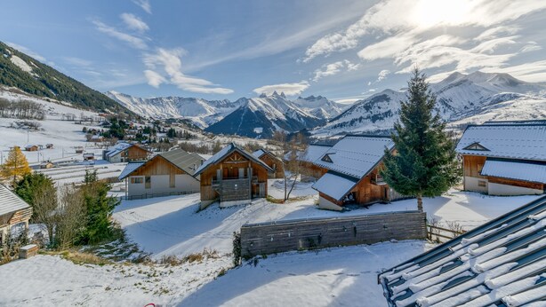 A snow-covered village with wooden cabins, a wooden fence, and a mountain backdrop.