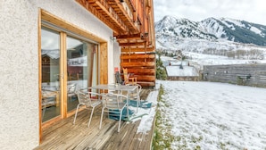 A wooden deck with a table and chairs, a glass door leading to a room, and a snowy mountain backdrop.