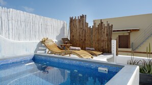 Een zwembad met blauwe tegels, houten lounge stoelen, een witte muur en een houten hek.