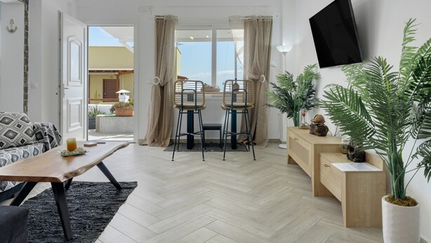 Een moderne woonkamer met een houten salontafel, twee barkrukken, een aan de muur bevestigde televisie en een potplant.