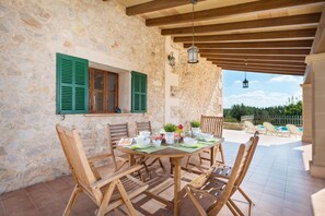 Un espace extérieur aménagé pour les repas, avec des murs en pierre, des chaises en bois et une table dressée, offrant une vue sur une piscine et la verdure environnante.