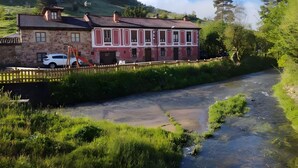 A stone house with a red and white facade, a wooden fence, and a river in the foreground.