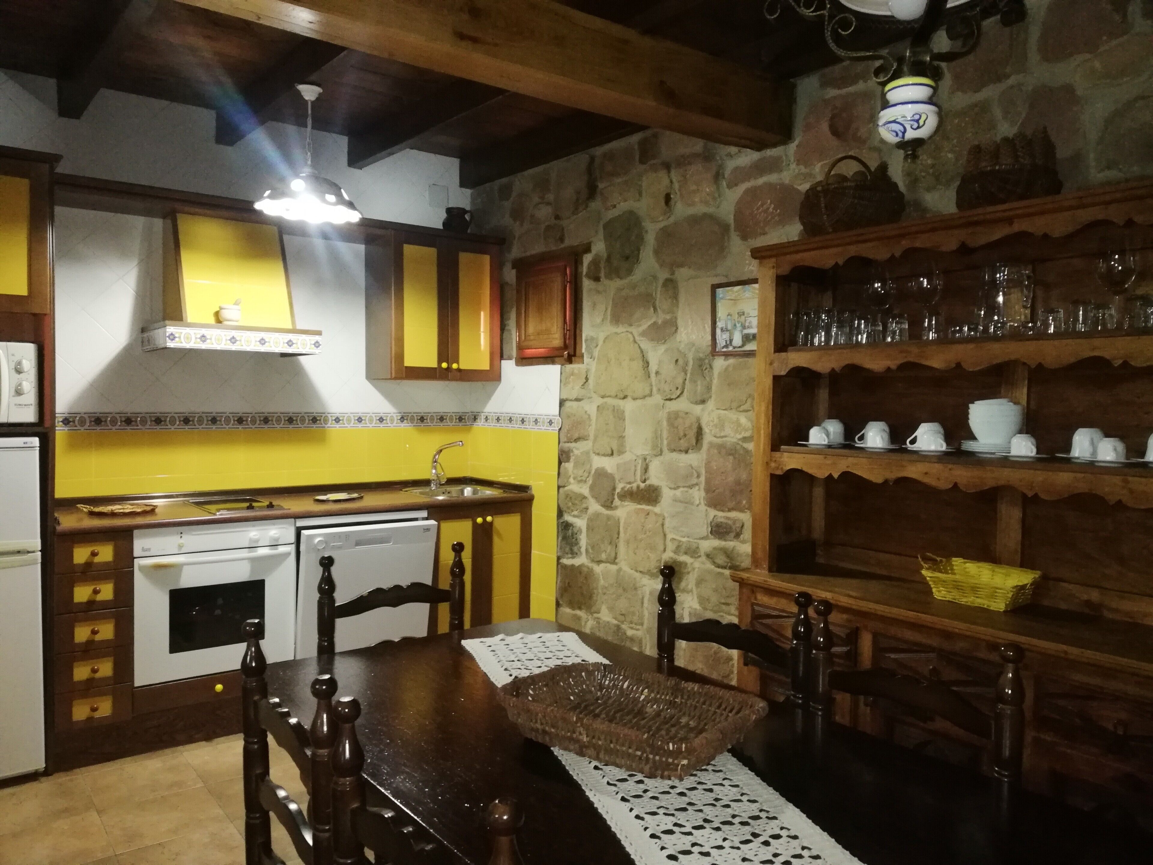 A kitchen with a stone wall, yellow cabinets, a wooden table, and a shelf with glassware.