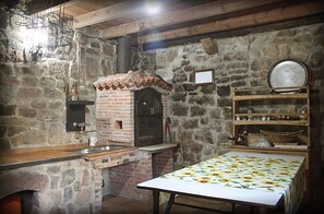 Une cuisine rustique avec un four en briques, des étagères en bois et une table recouverte d’une nappe à motifs floraux.