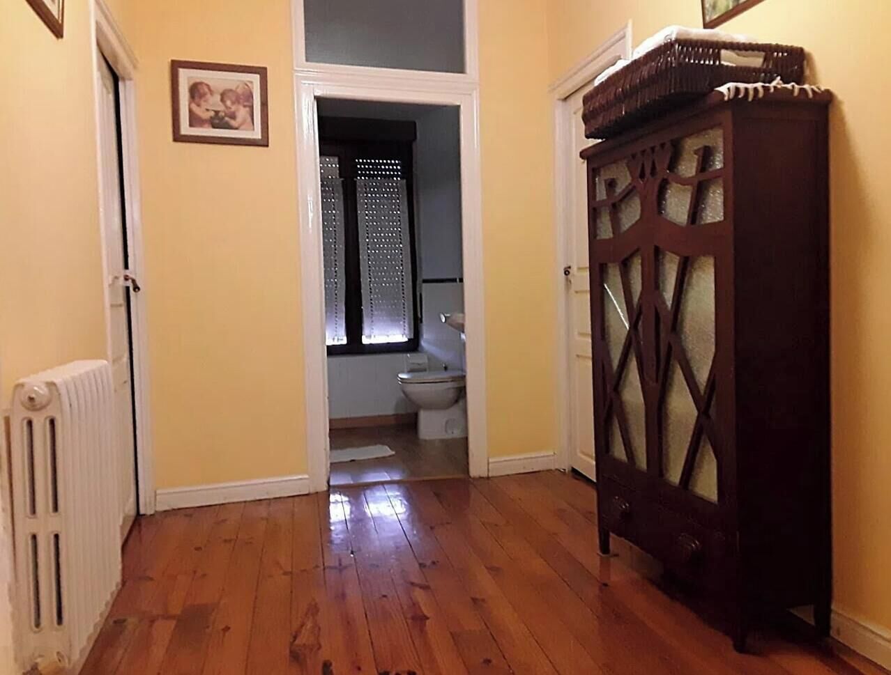 Ein Flur mit Holzboden, einem Heizkörper, einem dunklen Holzschrank und einer Tür zu einem Badezimmer mit Toilette und Fenster.