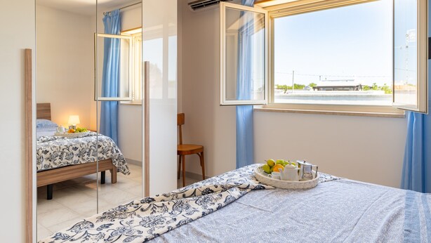 Ein Schlafzimmer mit Bett, Sessel, einem fensterbepflanzten Fenster und einem Spiegel, der den Raum reflektiert.