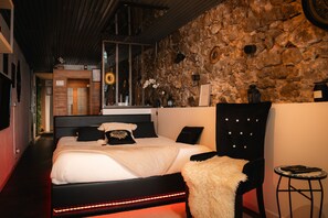 A bedroom with a stone wall, a bed with black and white bedding, a black chair, and a small table with a lamp.