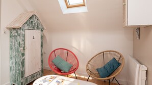 Une pièce avec un plafond en pente, une petite lucarne, un meuble en bois, deux fauteuils en osier et une table avec des magazines.