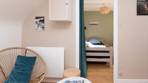 Une chambre à coucher comprenant un lit, une chaise en osier, une petite table ronde, une étagère fixée au mur et une porte donnant sur une autre pièce.