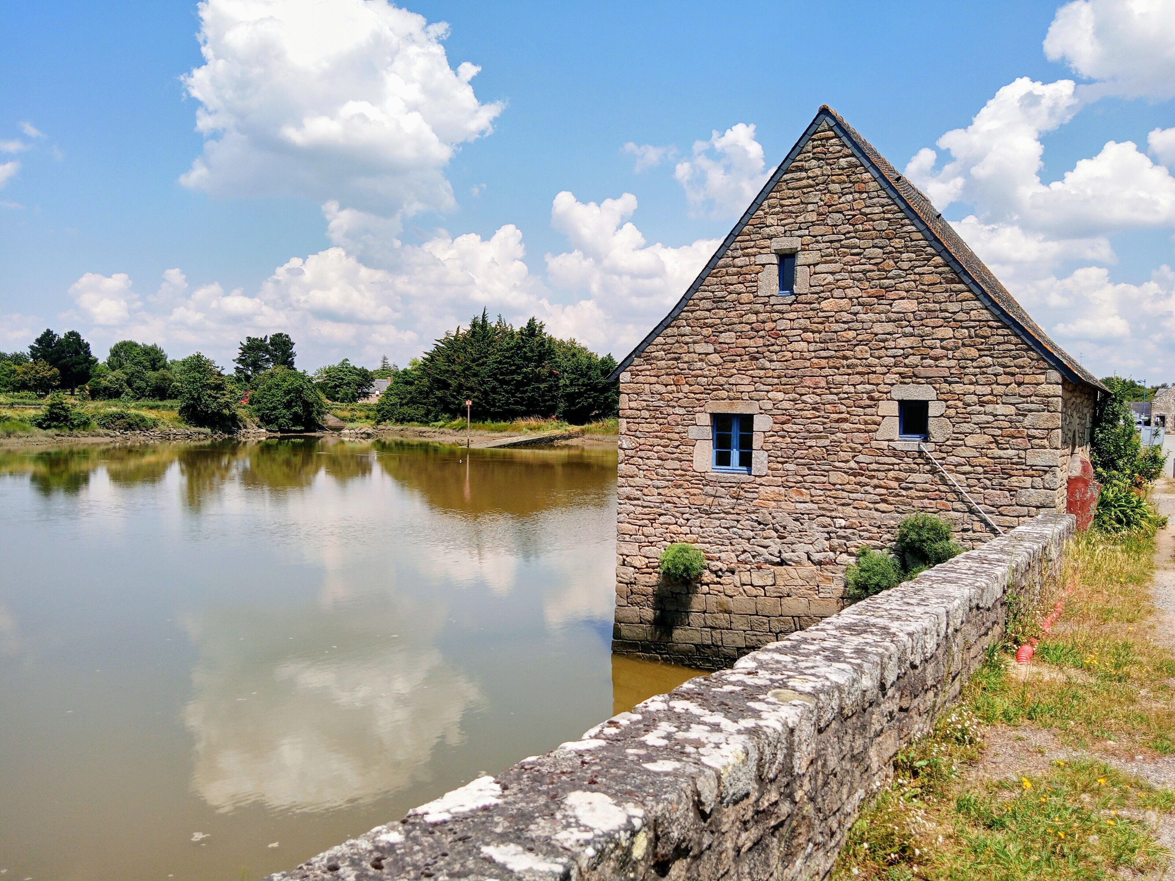 A stone building with a triangular roof by a calm body of water, surrounded by greenery.