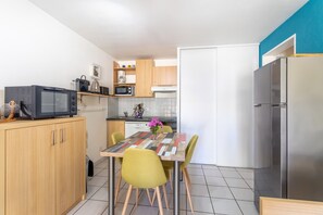 Cozinha compacta com micro-ondas, torradeira e frigorífico. Mesa de jantar com cadeiras e um vaso com flores.