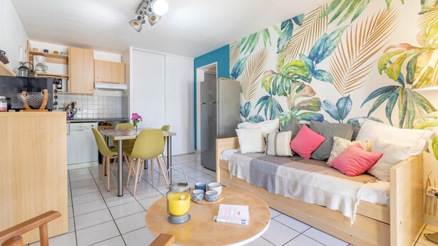 A modern kitchen with wooden cabinets, a dining area with a table and chairs, a living area with a sofa and cushions, and a tropical leaf-patterned wall. - Apartment 'T2 Meublé Aux Aloès' with Mountain View, Shared Pool and Private Terrace (Cerbère)