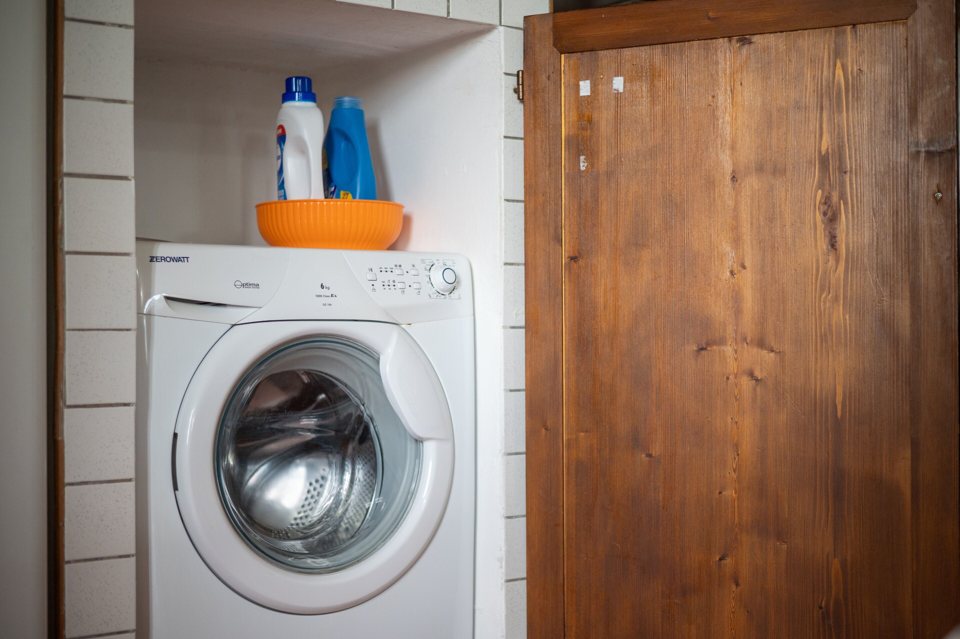 Una lavadora de carga frontal con panel de control, una puerta de madera y un estante con botellas de detergente.