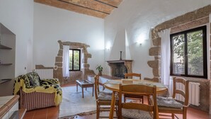 A rustic interior with exposed stone walls, a fireplace, a dining table with chairs, and a sofa with a patterned cover.