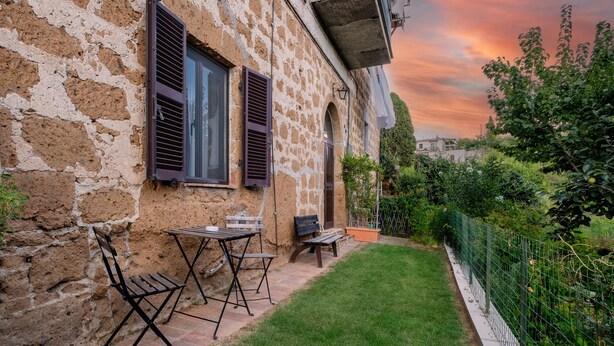 A rustic stone building with a small patio, outdoor furniture, and a green fence.