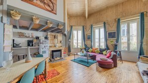 Amplia sala de estar con chimenea, pisos de madera, un sofá, una mesa de comedor con sillas y una zona de cocina con estantes y electrodomésticos.