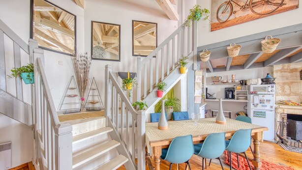 Una escalera con barandillas blancas, una mesa de comedor de madera con sillas azules, y una zona de cocina con microondas y refrigerador.