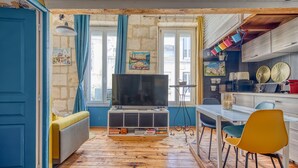 A room with a TV, wooden flooring, a sofa, a dining table with chairs, and a kitchen area. - Dari Apartment – Elegant Getaway in the Heart of Bordeaux (Bordeaux)