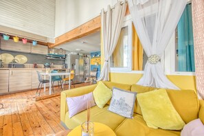 A modern living room with a yellow sofa, a dining area, and a kitchen with wooden beams.