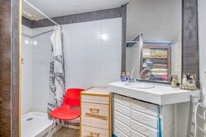 A bathroom with a shower, a red chair, a sink with decorative items, and a mirror reflecting a storage area.