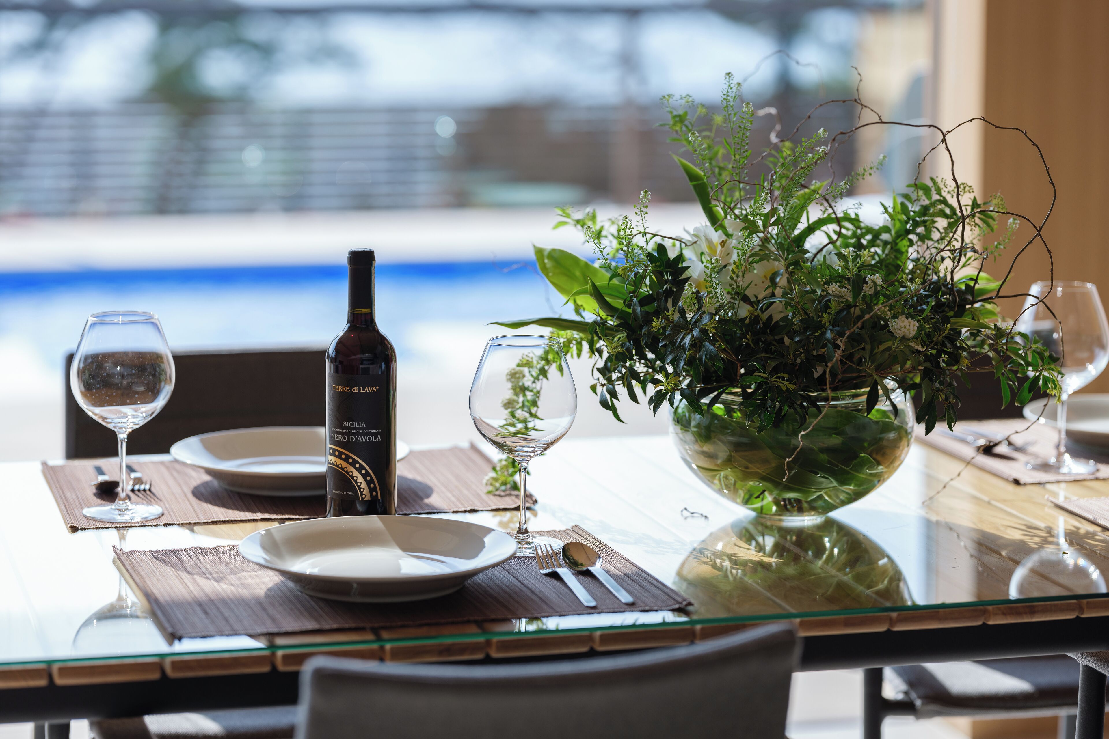 A dining table with a bottle of wine, a glass, a bowl of greenery, and a view of a pool.
