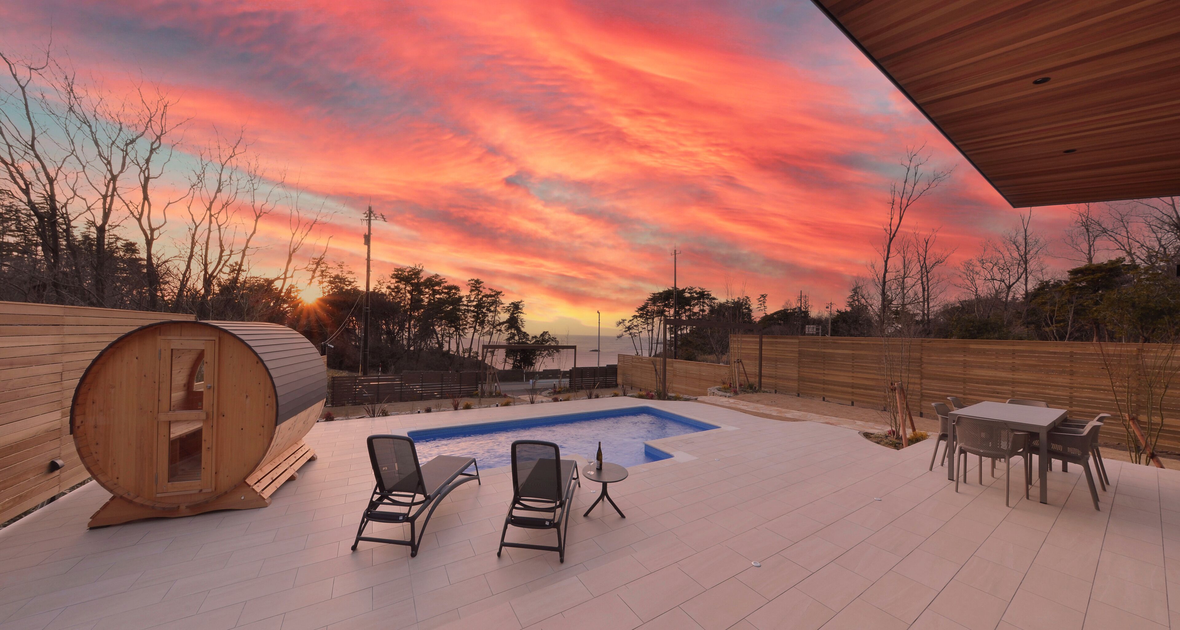 A wooden sauna, two lounge chairs, a small pool, and a table with chairs on a patio with a wooden fence and trees in the background.