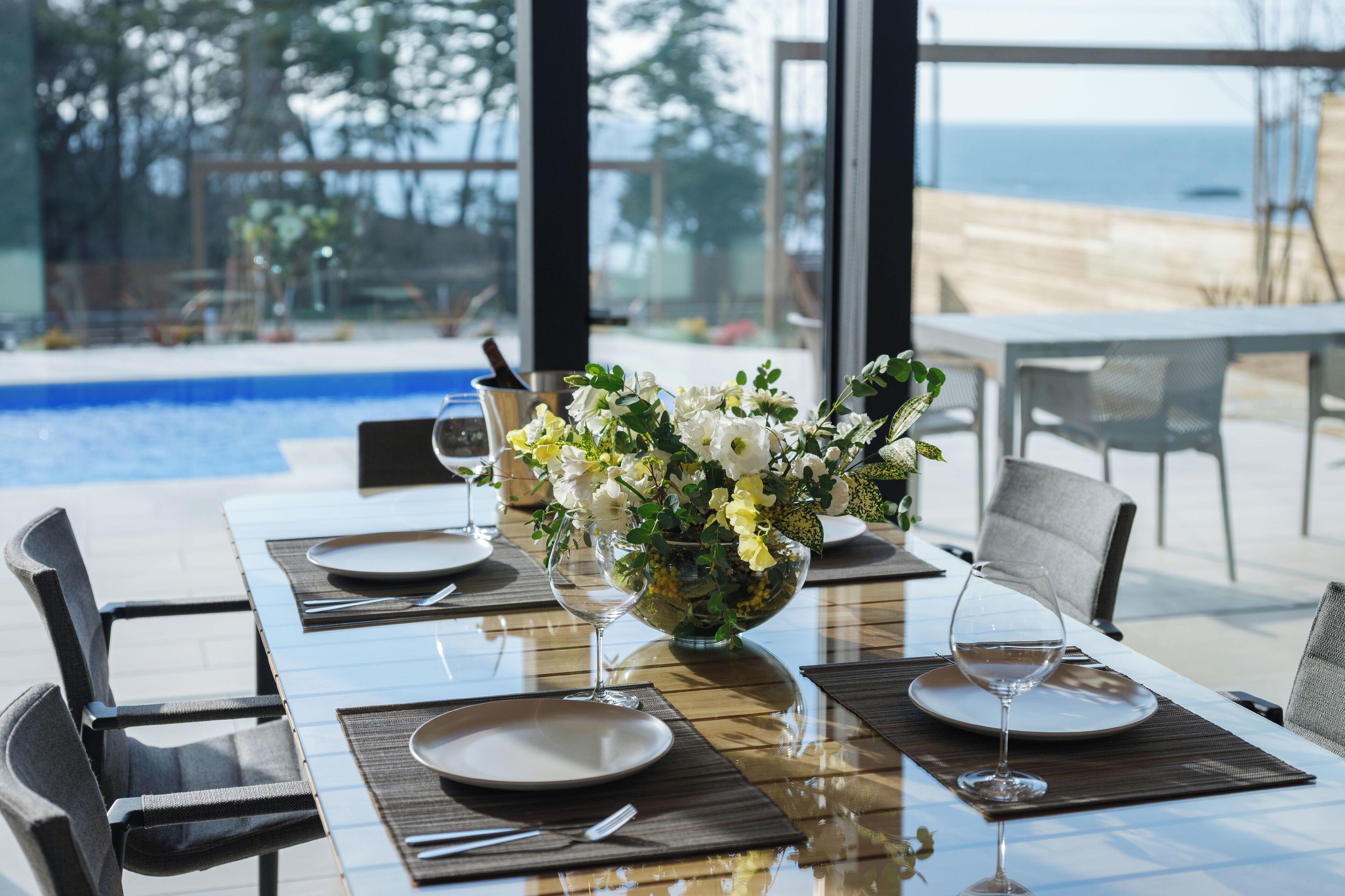 A dining table set for two with a vase of flowers, a glass of water, and a view of the ocean.