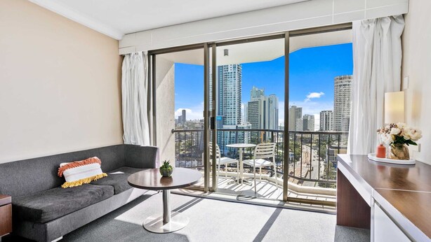 A modern hotel room with a grey sofa, a small round table, and a balcony with a city view. - Ocean View Surfers Paradise Studio Suite (Surfers Paradise)