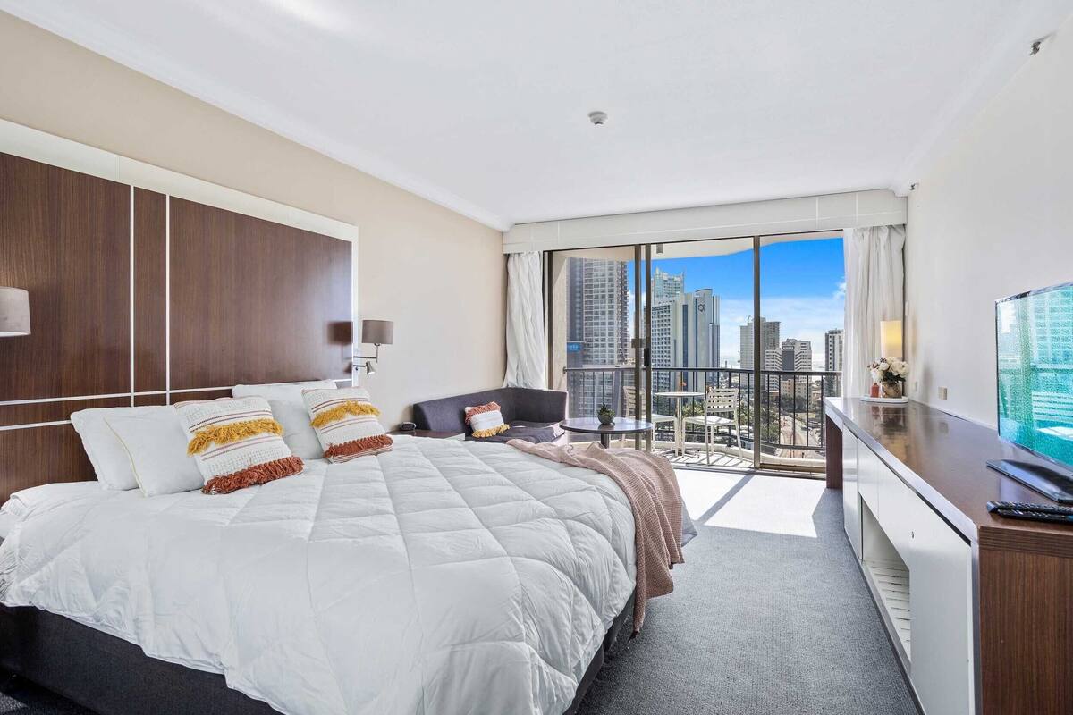 A modern hotel room with a large bed, a desk, and a balcony with a city view.